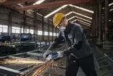 Seseorang yang mengenakan peralatan keselamatan menggiling logam, menghasilkan percikan terang di pabrik.