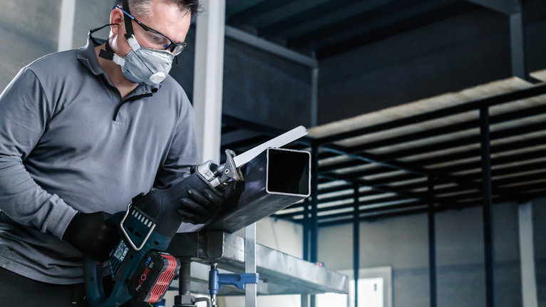 Seseorang yang mengenakan peralatan keselamatan menggunakan gergaji bolak-balik untuk memotong pipa logam.