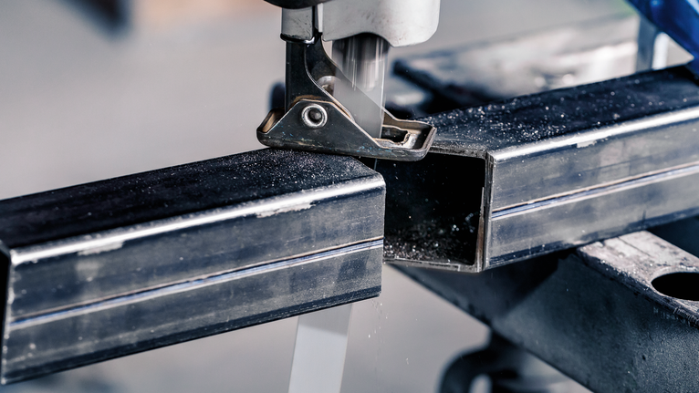 Memotong tabung persegi logam dengan gergaji ukir pada meja kerja.