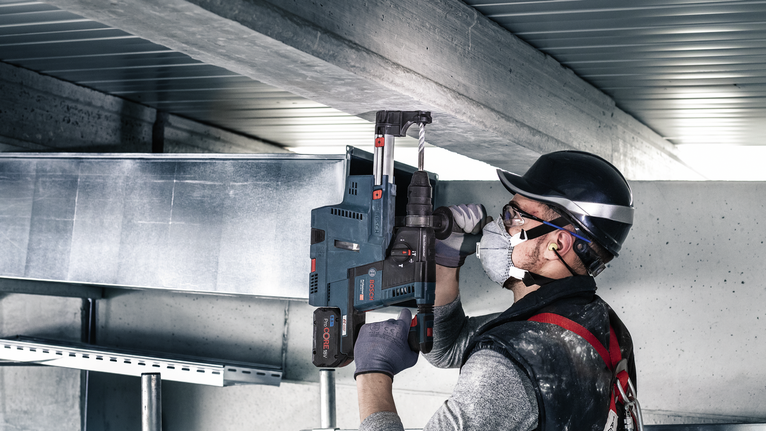 Seseorang yang mengenakan peralatan keselamatan mengebor balok logam di lokasi konstruksi.