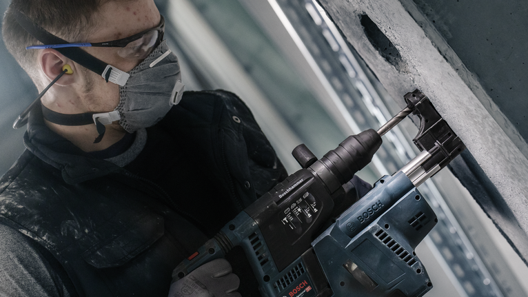 Seseorang yang mengenakan peralatan keselamatan mengebor dinding beton dengan alat listrik.