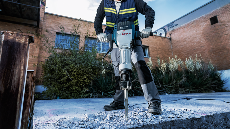 Seseorang yang mengenakan peralatan keselamatan memecahkan beton dengan palu pembongkaran di luar ruangan.