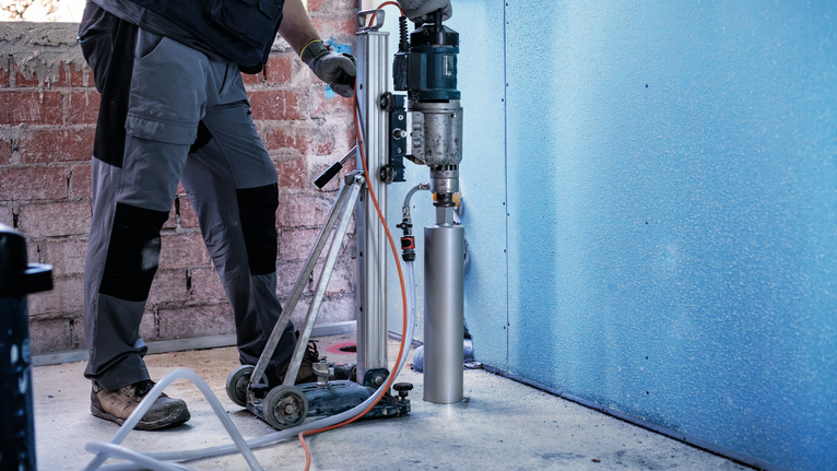 Seseorang yang mengenakan peralatan keselamatan mengebor beton dengan mesin pengebor inti yang terpasang.