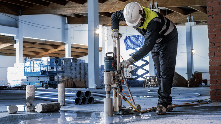 Seseorang yang mengenakan peralatan keselamatan mengebor beton di lokasi konstruksi.