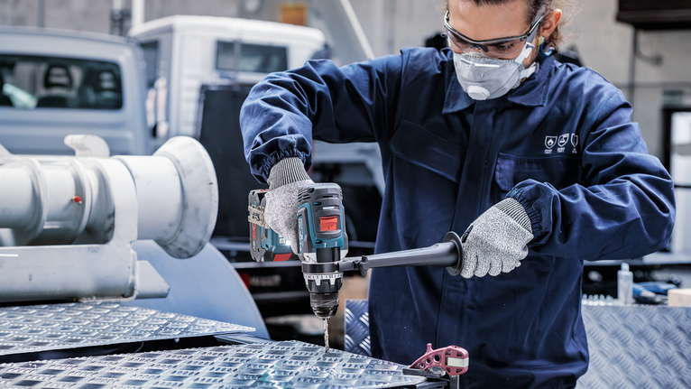 Orang yang mengenakan peralatan keselamatan mengebor pelat logam di lingkungan industri.