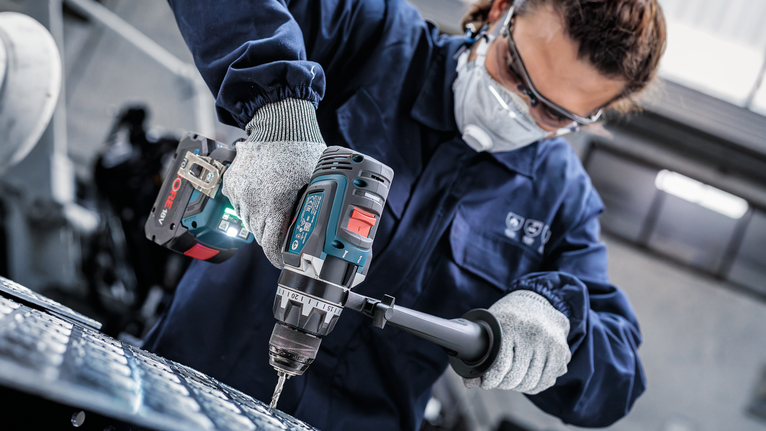 Orang yang mengenakan peralatan keselamatan mengebor logam dengan alat listrik tanpa kabel.