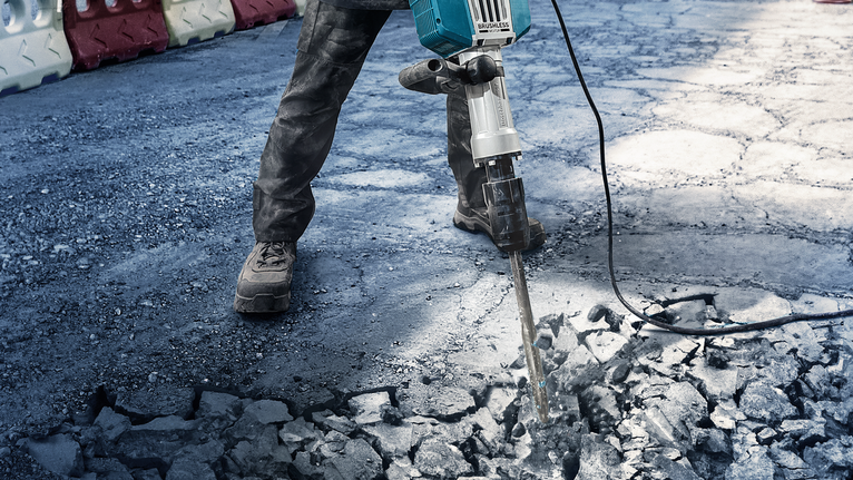 Seseorang yang mengenakan peralatan keselamatan memecahkan beton dengan alat pembongkaran tugas berat.