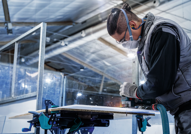 Orang yang mengenakan peralatan keselamatan memotong panel kayu dengan alat listrik.