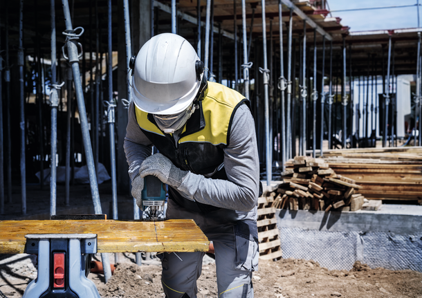 Seseorang yang mengenakan peralatan keselamatan memotong papan kayu dengan alat listrik di lokasi konstruksi.