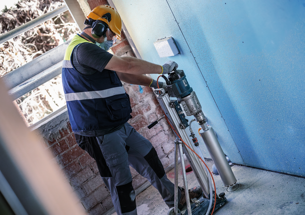 Seseorang yang mengenakan peralatan keselamatan mengebor dinding beton menggunakan bor inti yang terpasang.