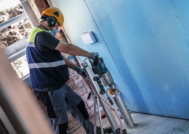 Seseorang yang mengenakan peralatan keselamatan mengebor dinding biru dengan alat listrik yang terpasang.