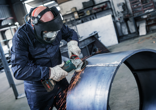 Orang yang mengenakan peralatan keselamatan menggiling pipa logam besar, sehingga menimbulkan percikan api.