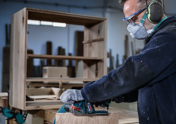 Orang yang mengenakan peralatan keselamatan mengampelas perabotan kayu dengan perkakas listrik.
