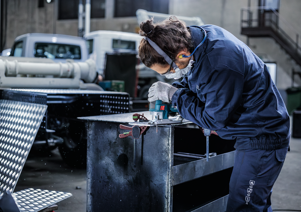 Seseorang yang mengenakan peralatan keselamatan mengebor logam di meja kerja di bengkel industri.