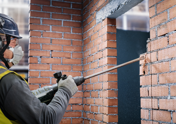 Orang yang mengenakan peralatan keselamatan memahat dinding bata dengan palu pembongkaran.