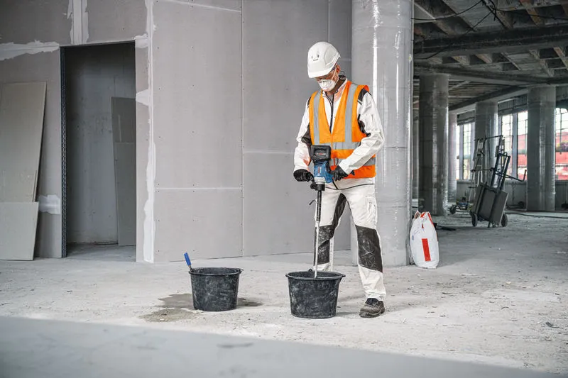 Seseorang yang mengenakan perlengkapan keselamatan menggunakan mekanisme pengadukan untuk mencampur material dalam ember.