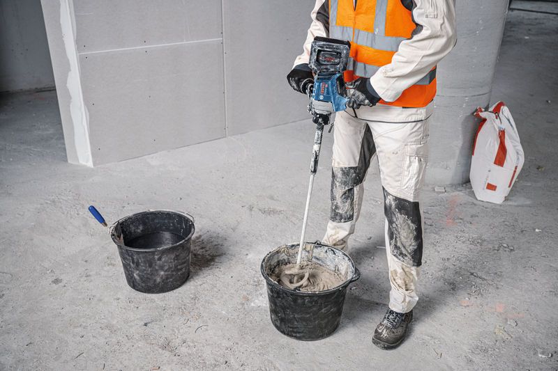 Seseorang yang mengenakan peralatan keselamatan mencampur material konstruksi dengan mekanisme pengadukan.