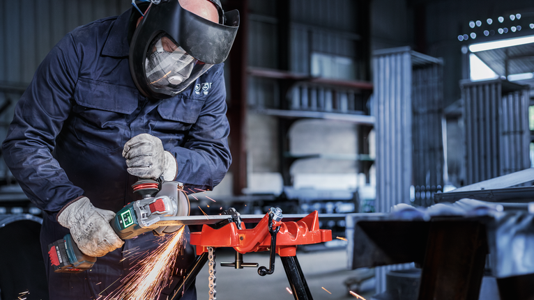 Orang yang mengenakan peralatan keselamatan menggunakan alat listrik untuk menggiling logam, sehingga menimbulkan percikan api.