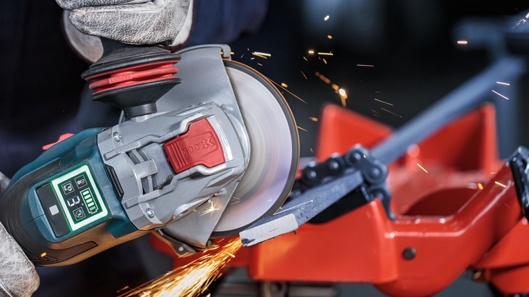 Orang yang mengenakan peralatan keselamatan menggunakan penggiling sudut untuk memotong batang logam.