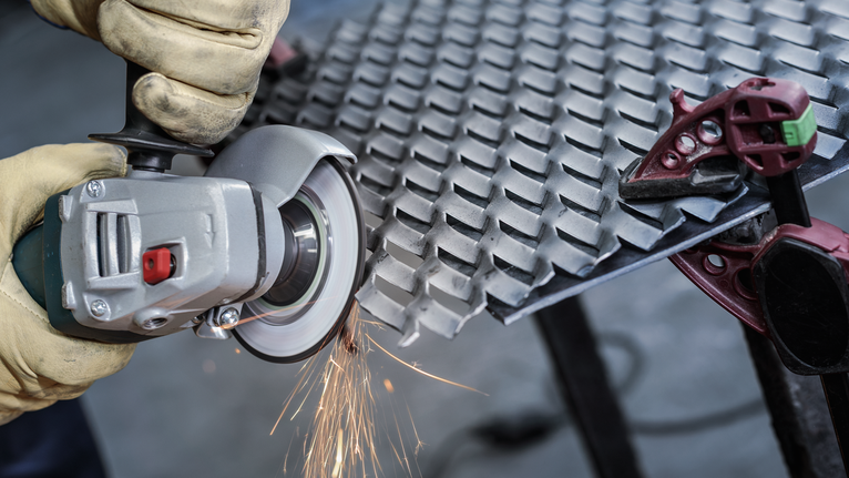 Orang yang mengenakan peralatan keselamatan menggunakan penggiling sudut untuk memotong lembaran logam.