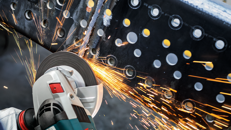 Orang yang mengenakan peralatan keselamatan menggiling balok logam dengan alat listrik, sehingga menimbulkan percikan api.