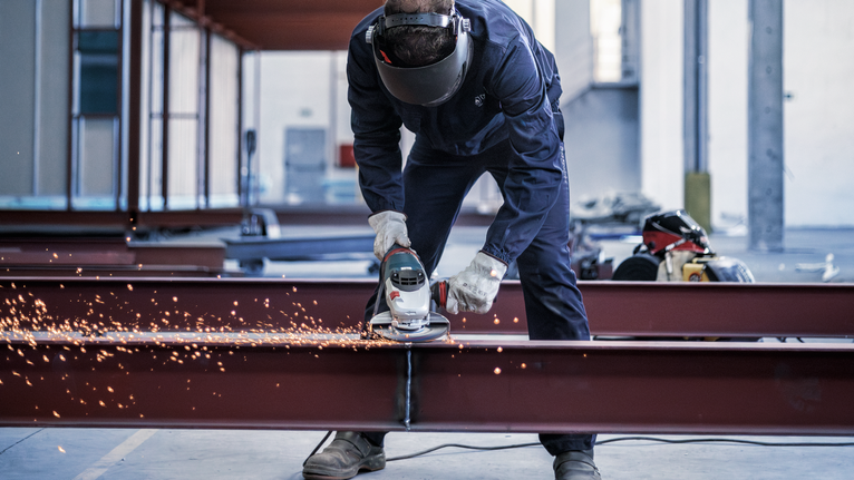 Orang yang mengenakan peralatan keselamatan menggiling balok baja, menghasilkan percikan terang.