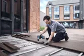Seseorang yang mengenakan peralatan keselamatan menggunakan bor tanpa kabel untuk memasang papan dek di luar.