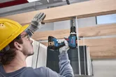 Seseorang yang mengenakan peralatan keselamatan mengebor sekrup ke balok kayu.