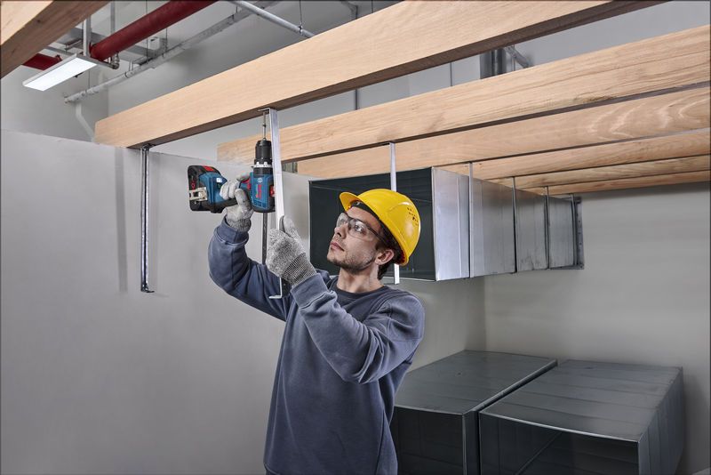 Seseorang yang mengenakan peralatan keselamatan mengebor batang logam di bawah balok kayu.