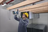 Seseorang yang mengenakan peralatan keselamatan mengebor batang logam di bawah balok kayu.