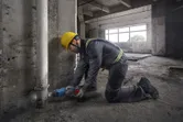 Seseorang yang mengenakan peralatan keselamatan menggunakan gergaji bolak-balik untuk memotong pipa di dekat dinding beton.
