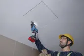 Seseorang yang mengenakan peralatan keselamatan memotong panel langit-langit dengan gergaji bolak-balik.