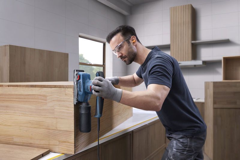 Seseorang yang mengenakan perlengkapan keselamatan sedang mengampelas lemari kayu menggunakan sander orbital.