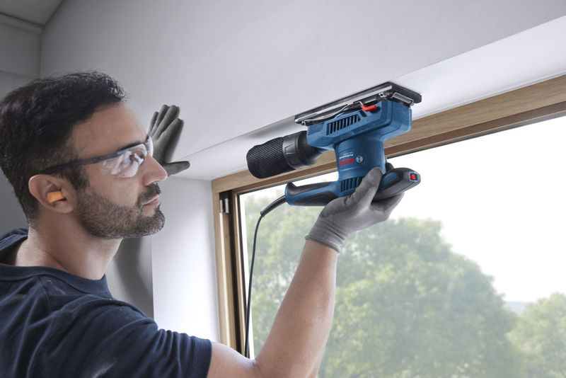 Seseorang yang mengenakan perlengkapan keselamatan mengampelas bingkai jendela dengan sander orbital.