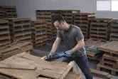 Seseorang yang mengenakan peralatan keselamatan memotong palet kayu dengan gergaji bolak-balik.