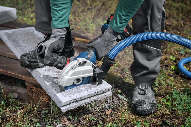 Seseorang yang mengenakan peralatan keselamatan menggunakan penggiling sudut besar dengan penyedot debu untuk memotong beton.