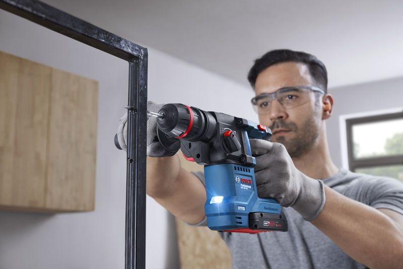 Seseorang yang mengenakan perlengkapan keselamatan mengebor rangka logam menggunakan palu putar tanpa kabel.