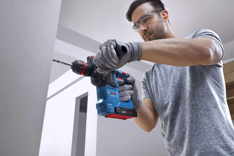 Seseorang yang mengenakan peralatan keselamatan mengebor dinding beton menggunakan palu putar tanpa kabel.