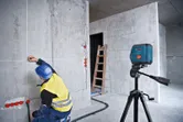 Seseorang yang mengenakan perlengkapan keselamatan menandai dinding menggunakan alat perataan laser.