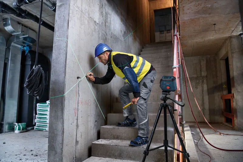 Seseorang yang mengenakan perlengkapan keselamatan menandai dinding beton menggunakan alat perataan laser.
