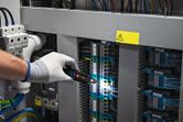 Seseorang yang mengenakan peralatan keselamatan memeriksa kabel listrik di panel kontrol dengan penguji tegangan.