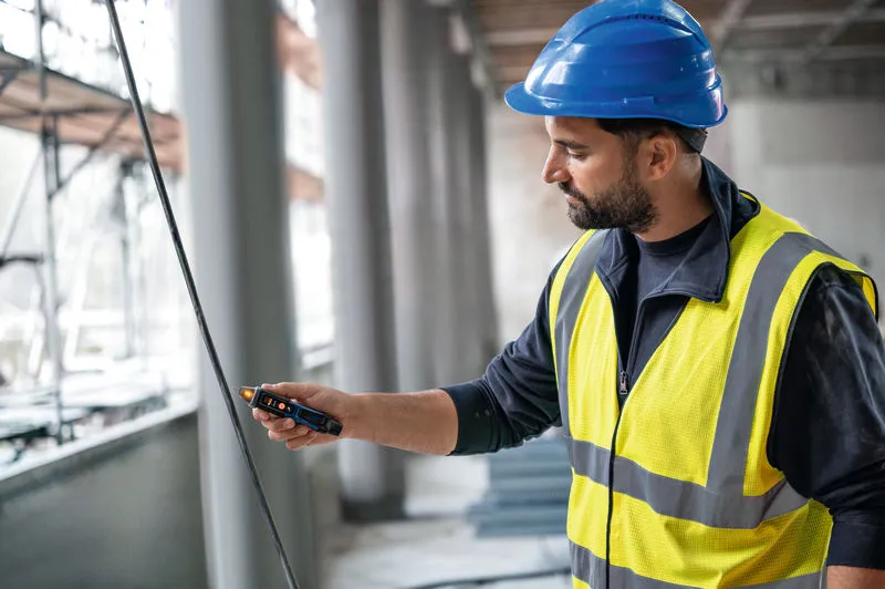 Seseorang yang mengenakan peralatan keselamatan menguji kabel dengan penguji tegangan non-kontak.