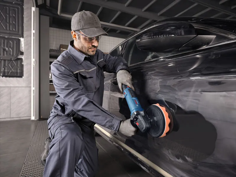 Seseorang yang mengenakan perlengkapan keselamatan memoles mobil hitam menggunakan perkakas listrik.