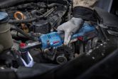 Seseorang yang mengenakan perlengkapan keselamatan menggunakan kunci ratchet tanpa kabel untuk mengencangkan baut di mesin mobil.