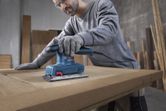 Seseorang yang mengenakan perlengkapan keselamatan mengampelas pintu kayu dengan sander orbital.