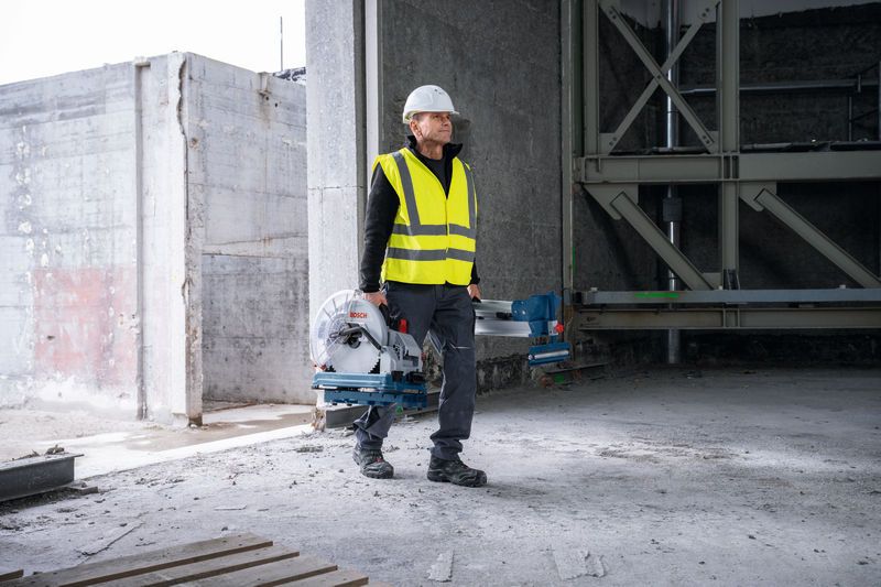 Seseorang yang mengenakan peralatan keselamatan membawa gergaji pemotong logam di sebuah bangunan yang belum selesai.