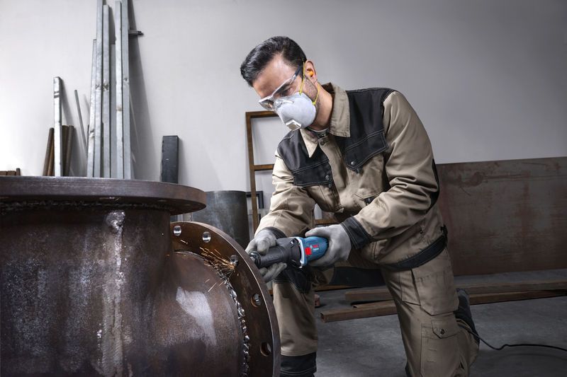 Seseorang yang mengenakan perlengkapan keselamatan menggerus tepian pipa logam besar.