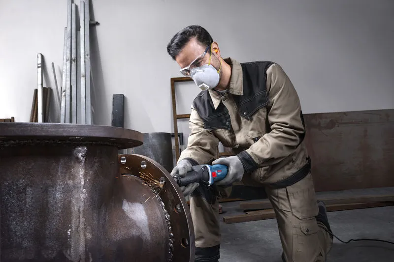 Seseorang yang mengenakan perlengkapan keselamatan menggerus tepian pipa logam besar.