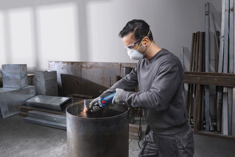 Seseorang yang mengenakan peralatan keselamatan mengasah tepi tong logam dengan penggiling lurus.