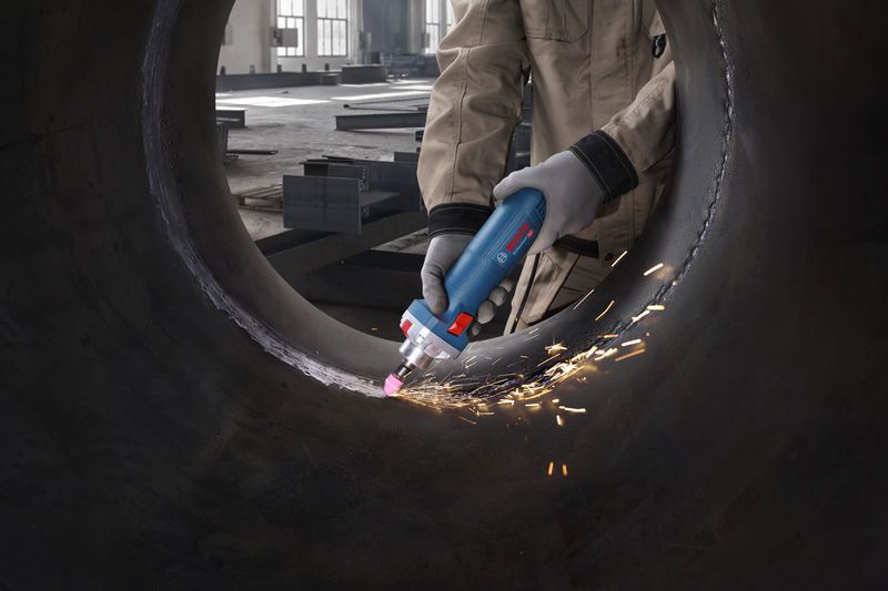 Seseorang yang mengenakan peralatan keselamatan menggiling bagian dalam pipa logam besar dengan penggiling lurus.